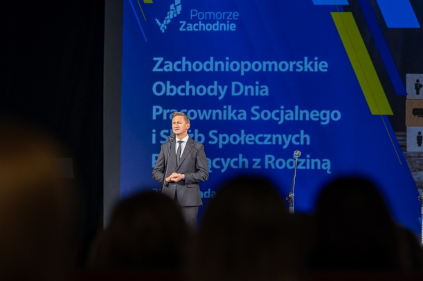 marszałek województwa Olgierd Geblewicz, przed marszałkiem siedzi publiczność, goście uroczystości, w tle niebieski napis z nazwą uroczystości