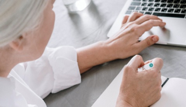 kobieta o siwych włosach zapisująca na kartkach notesu dane z laptopa